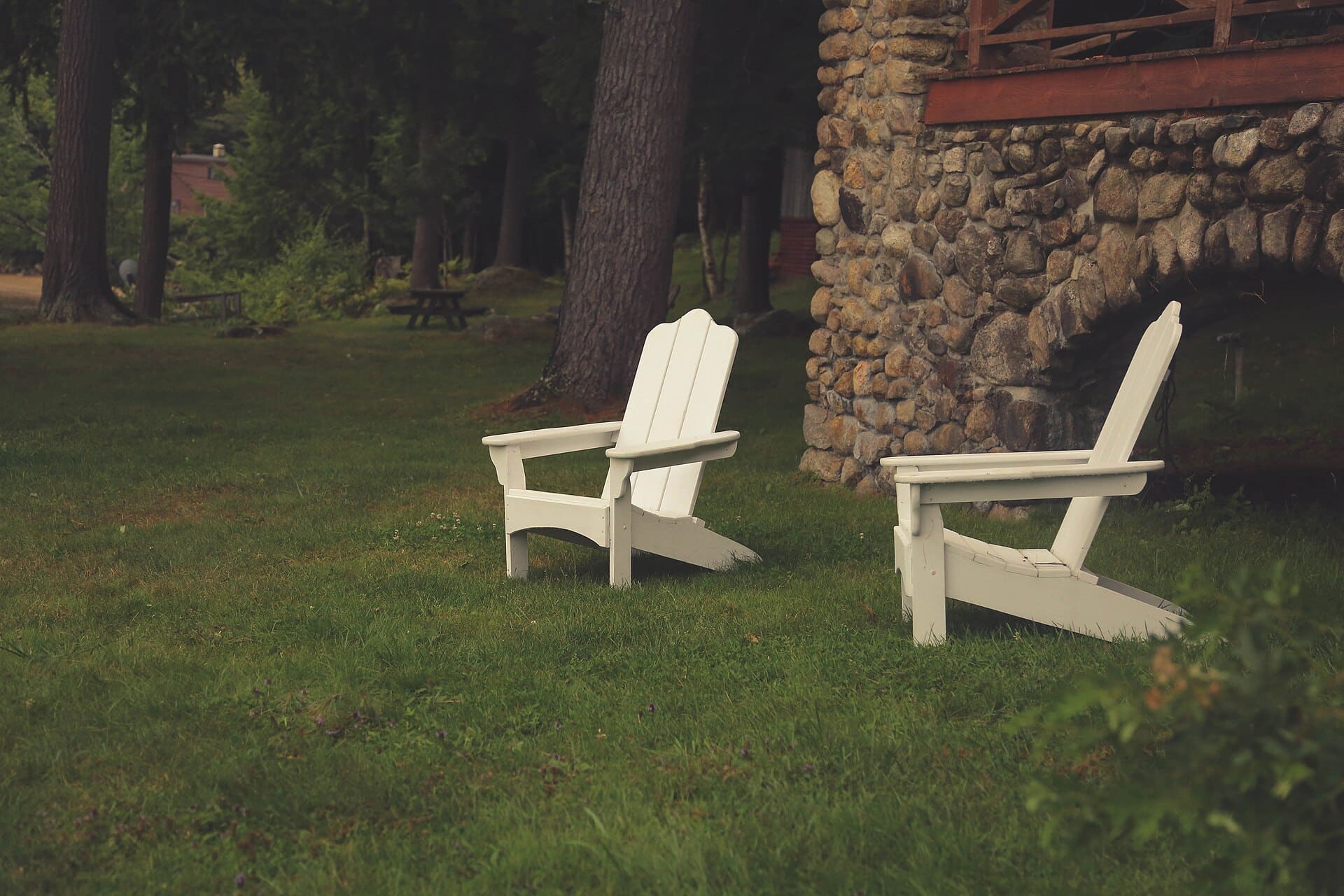 Two white Adirondack chairs turned toward each other on a grassy lawn beside a stone cabin and tall pine trees